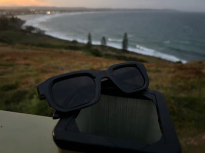 Black sunglasses on a surface with a scenic view of a beach and lighthouse in the background.