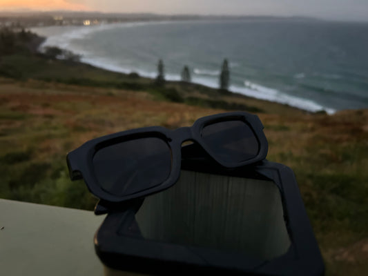 Black sunglasses on a surface with a scenic view of a beach and lighthouse in the background.